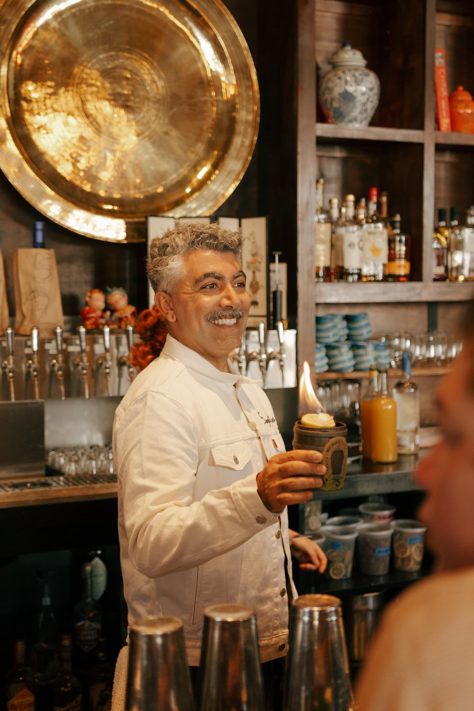 A bartender holding a flaming drink beneath a brass tray