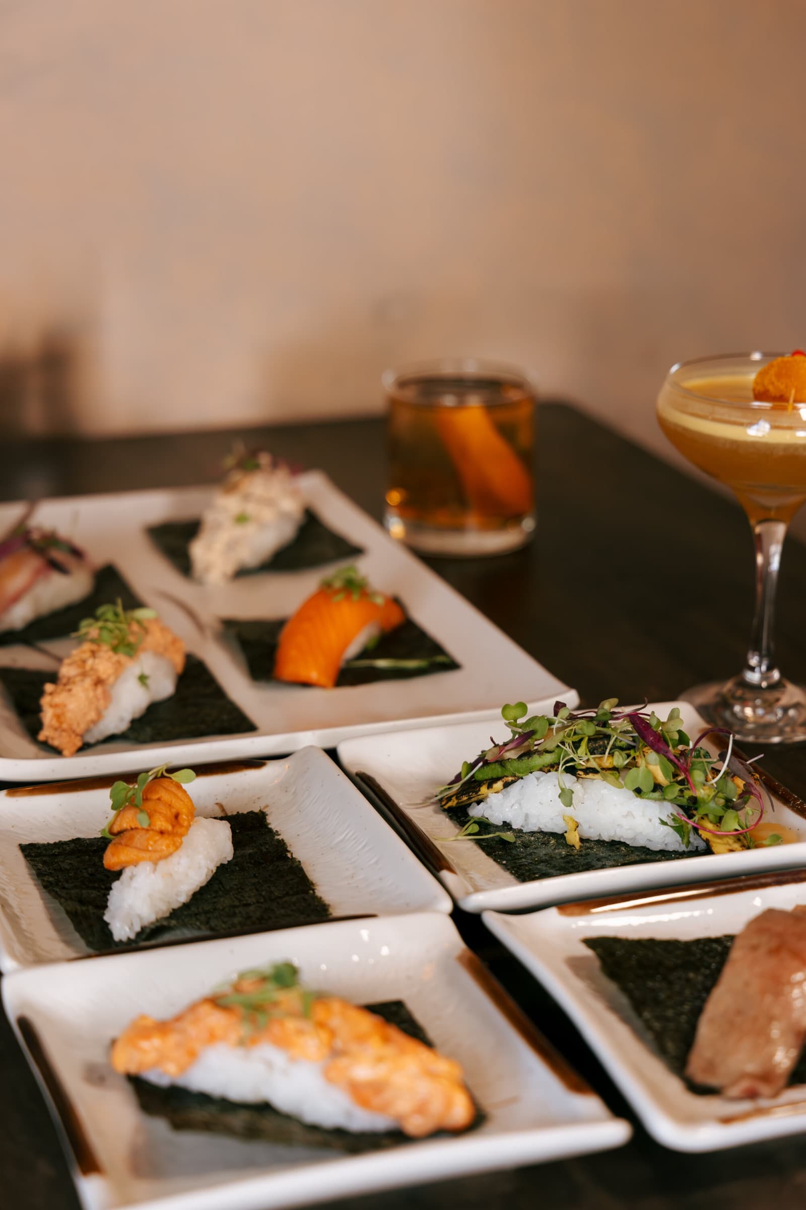 Nigiri arranged on small plates, with cocktails behind