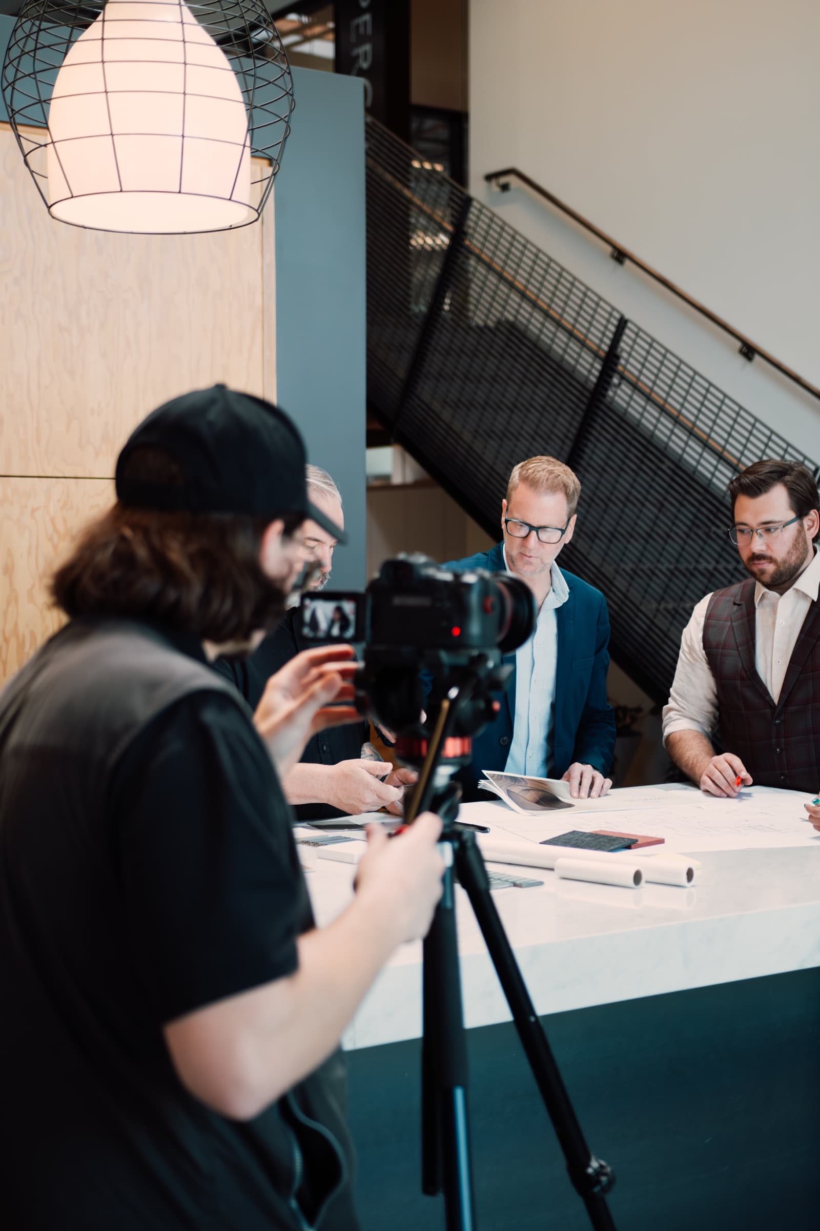A camera operator filming three men reviewing drawings at a table