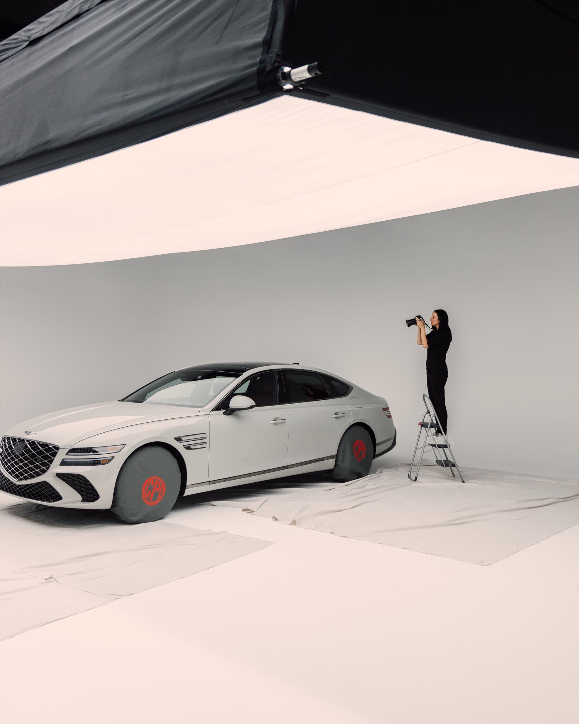 A photographer on a ladder shooting a white sedan in a studio